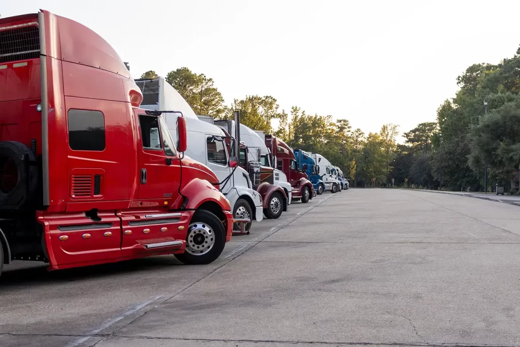 A variety of semi-trucks aligned at an angle with green trees in the background at dawn.