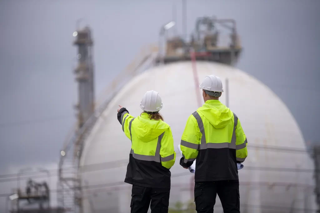 Engineers wearing safety gear, including hard hats examining survey a large blueprint tablet standing industrial facility gas or oil refinery engaged in a job requires high safety standards concept.